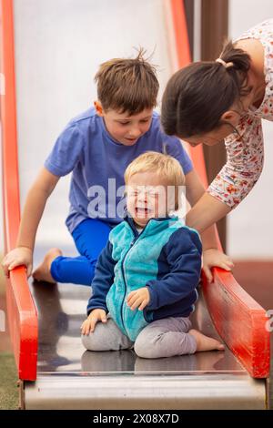 Eine pflegende Familie hilft ihrem Baby mit Down-Syndrom auf einem Spielplatz, um Sicherheit zu gewährleisten und auf einer Rutsche Unterstützung zu bieten. Stockfoto