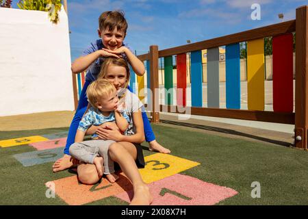 Fröhliche Familie mit einem kleinen Jungen und einem Baby mit Down-Syndrom, die an einem sonnigen Tag auf einem farbenfrohen Spielplatz Zeit verbringen. Stockfoto