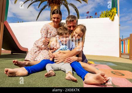 Eine glückliche Familie genießt einen verspielten Moment zusammen auf einem Spielplatz im Freien. Die Inclusive-Szene zeigt ein kleines Baby mit liebevoller Embra des Down-Syndroms Stockfoto