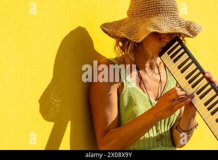 Eine Straßenmusikerin mit großem Strohhut spielt eine Melodika vor einer leuchtend gelben Wand und wirft einen Schatten in der sonnigen Atmosphäre. Stockfoto