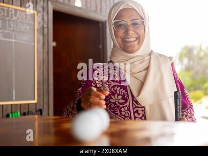 Fröhliche muslimische Frau im Hijab, die in die Kamera schaut, während sie Minigolf spielt, um einen Golfball zu nehmen. Stockfoto