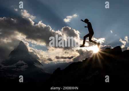 Dieses Bild, das bei Sonnenuntergang aufgenommen wurde, zeigt eine Person, die sich mitten im Sprung gegen die Sonnenstrahlen befindet, und das Matterhorn bietet eine atemberaubende Kulisse Stockfoto