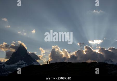 Zwei Wanderer stehen vor einem Himmel, wo Sonnenstrahlen in der Abenddämmerung durch die Wolken über dem Matterhorn brechen und die Schönheit des Schweizer Al hervorheben Stockfoto