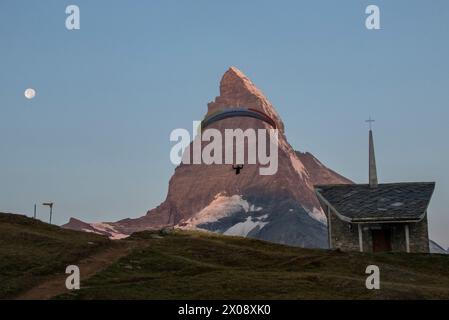 Ein Gleitschirmflieger fliegt in der Nähe des berühmten Matterhorns, während der Mond am Himmel hängt, mit einer malerischen Kapelle im Vordergrund Stockfoto