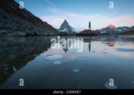 Eine ruhige Frühlingsszene fängt einen teilweise aufgetauten See ein, der das berühmte Matterhorn in den Schweizer Alpen reflektiert, mit einer nicht erkennbaren Person in s Stockfoto