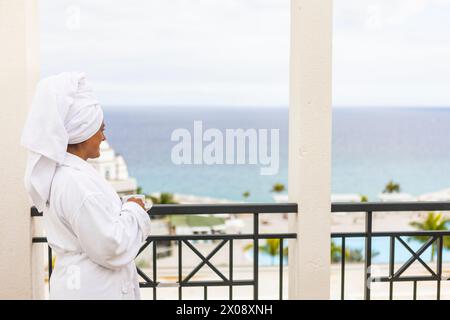 Rückansicht einer hispanischen Frau mittleren Alters, die in einen weißen Bademantel mit einem Handtuch auf dem Kopf gewickelt ist, genießt eine Tasse Kaffee auf einem Balkon mit Blick auf das Meer Stockfoto