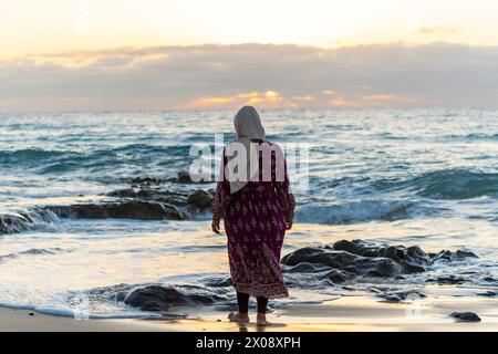 Eine muslimische Frau steht kontemplativ an der Küste und blickt auf einen ruhigen Sonnenuntergang im Ozean, mit Wellen, die sanft die Felsen und den Sand streicheln Stockfoto