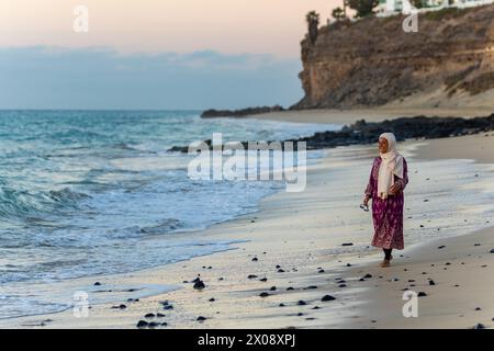 Eine muslimische Frau genießt einen friedlichen Spaziergang entlang der Küste, während ihr traditioneller Hijab und ihre Kleidung in Kontrast zur natürlichen Strandlage in der Dämmerung stehen Stockfoto