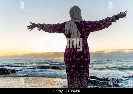 Muslimische Frau im Hijab genießt die Freiheit am Strand bei Sonnenuntergang, mit weit geöffneten Armen Stockfoto