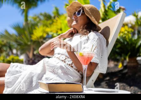 Eine hispanische Frau mittleren Alters genießt einen sonnigen Tag im Freien mit einem erfrischenden Cocktail und einem offenen Buch, das Ruhe und Freizeit ausstrahlt. Stockfoto