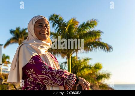 Eine fröhliche muslimische Frau, die einen weißen Hijab und ein gemustertes Kleid trägt, lächelt warm, während sie am Meer unter einem klaren blauen Himmel steht Stockfoto