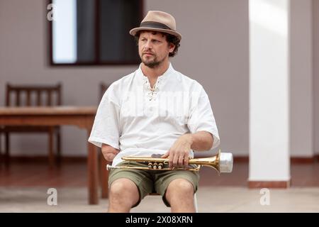 Ein Mann sitzt draußen auf einem Hocker, hält eine Trompete und trägt legere Kleidung, die ein entspanntes Ambiente mit musikalischem Thema weckt Stockfoto
