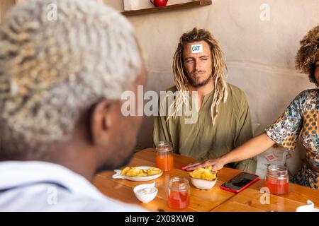 Eine vielfältige Gruppe von Freunden genießt ein lustiges Ratespiel mit Haftnotizen in einer gemütlichen Bar, wo sie Lachen und Snacks teilen Stockfoto