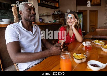 Zwei Freunde, die ein Rätsel mit Wortkarten auf der Stirn spielen und in einer rustikalen Bar mit Snacks und Getränken sitzen, lachen Stockfoto