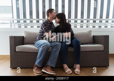Eine asiatische Frau und ein kaukasischer Mann umarmen und küssen die Stirn auf einem Sofa und zeigen Liebe und Begleitung über die Kulturen hinweg Stockfoto
