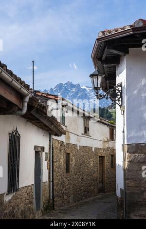 Traditionelle Steinhäuser säumen eine enge Straße in Potes, während im Hintergrund die majestätischen Picos de Europa unter einem klaren blauen Himmel thronen. Stockfoto