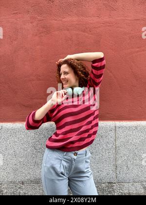 Lächelnde junge rothaarige Frau in lässiger Kleidung mit Kopfhörern um den Hals und posiert selbstbewusst vor einem leuchtend roten Hintergrund Stockfoto