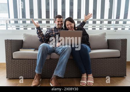 Ein fröhliches, multiethnisches Paar feiert spannende Neuigkeiten auf einem Laptop, während es zusammen auf einem Sofa sitzt und einen fröhlichen Moment teilt. Stockfoto