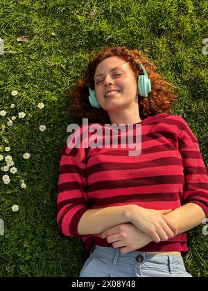 Eine junge rothaarige Frau genießt einen friedlichen Moment auf Gras mit Kopfhörern, umgeben von Wildblumen Stockfoto