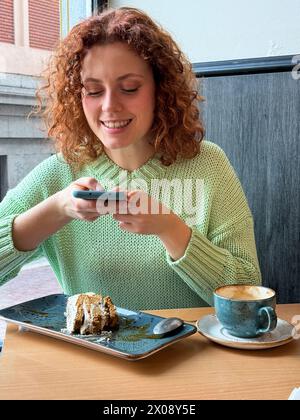 Eine fröhliche rothaarige Frau sitzt an einem Café-Tisch und macht Fotos mit ihrem Smartphone, mit einem teilweise gegessen Stück Kuchen und einer Tasse Kaffee Stockfoto