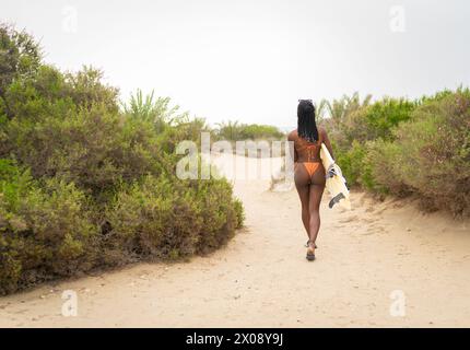 Die anonyme schwarze Frau mit einem Surfbrett geht auf einem Sandweg, umgeben von natürlichen Sträuchern, und weckt ein Gefühl von Sommerabenteuer und Entspannung Stockfoto