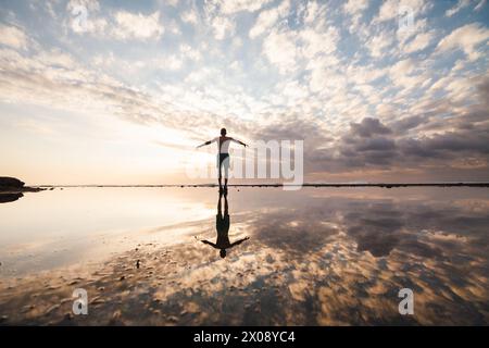 Silhouette einer Person mit ausgestreckten Armen an einem reflektierenden Strand bei Sonnenuntergang, unter einem bewölkten Himmel in Indonesien Stockfoto