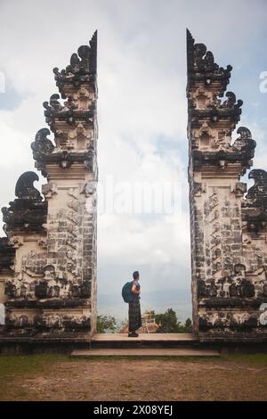 Ein Reisender steht zwischen den traditionellen geteilten Toren, Candi Bentar von Bali, symbolisiert den Eingang zum Heiligen, gekleidet in lokaler Kleidung und Cheri Stockfoto