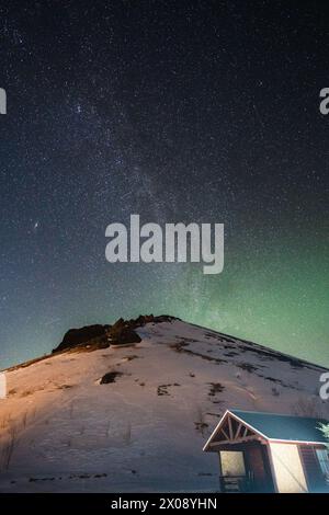 Ein atemberaubender Blick auf den sternenbesetzten Himmel und die subtilen Nordlichter über einem verschneiten Hügel, neben einer malerischen isländischen Hütte Stockfoto