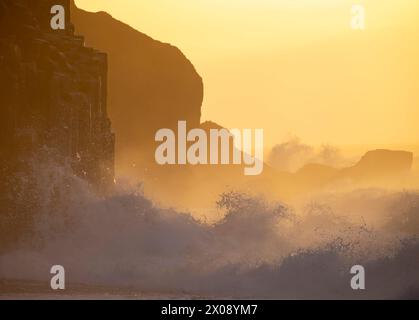 Das warme Leuchten des Sonnenuntergangs taucht die raue isländische Küste auf, während mächtige Wellen gegen Klippen ausbrechen und Sprühnebel in die Luft werfen Stockfoto