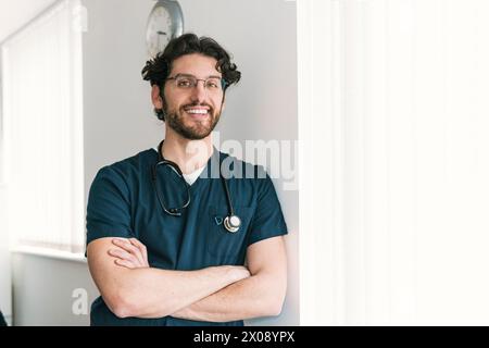 Ein nachdenklicher männlicher Gesundheitsarbeiter mit überkreuzten Armen steht an einer Uhr und symbolisiert rechtzeitige medizinische Dienste Stockfoto