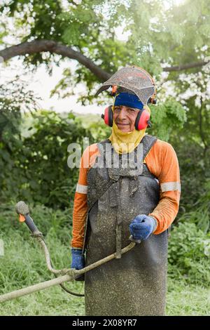 Ein erfahrener Landschaftsgärtner in Sicherheitsausrüstung hält einen Faden-Trimmer für ein Foto mitten im Grünen Stockfoto