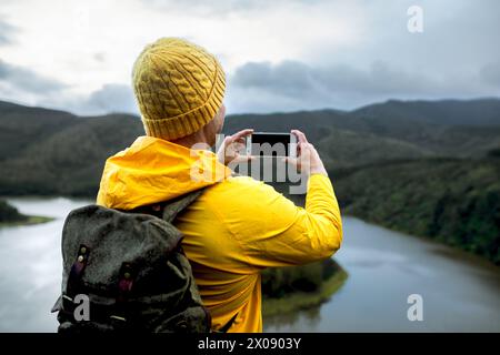 Rückansicht eines Mannes in einer gelben Jacke und Beanie steht mit Blick auf einen sich windenden Fluss und hält die Szene im Winter auf einem Smartphone fest. Stockfoto