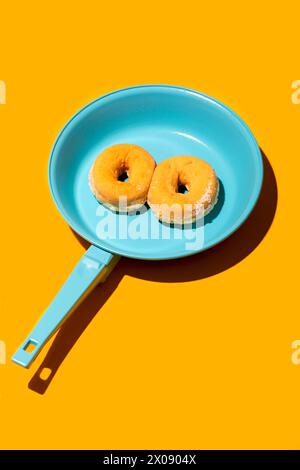 Ein lebhaftes Bild mit zwei goldenen gezuckerten Donuts, die auf einer hellblauen Pfanne vor einem leuchtend gelben Hintergrund liegen. Stockfoto