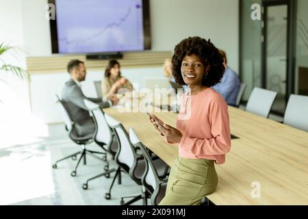 Ccheerful Afroamerikanerin leitet ein Meeting und hält sich an einem digitalen Tablet fest, während Kollegen hinter ihr Gespräche führen. Stockfoto