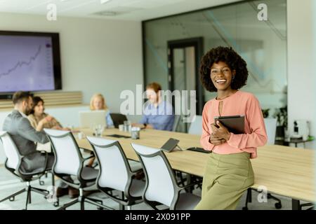 Ccheerful Afroamerikanerin leitet ein Meeting und hält sich an einem digitalen Tablet fest, während Kollegen hinter ihr Gespräche führen. Stockfoto