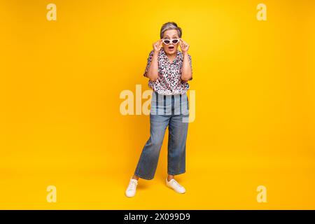 Foto in voller Länge der schönen älteren Dame schockiert nehmen Sie die Sonnenbrille gekleidet stilvolle Leopardendruck Kleid isoliert auf gelbem Hintergrund Stockfoto