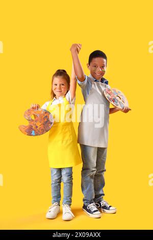 Niedliche kleine Kinder mit Gesichtern in Farben, die Hände auf gelbem Hintergrund halten Stockfoto