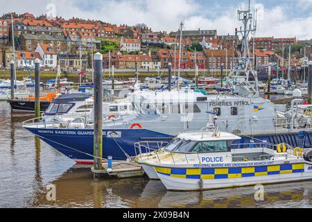 Ein Fischereipatrouillenboot und ein Polizeischiff liegen im Yachthafen von Whitby. Gebäude befinden sich in der Ferne auf einem Hügel und ein wolkengefüllter Himmel ist darüber. Stockfoto