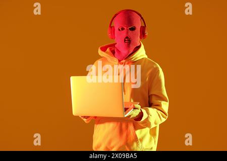 Schöner junger Mann in Sturmhaube mit Kopfhörern und Laptop auf gelbem Hintergrund Stockfoto