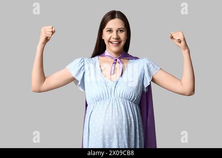 Schöne junge schwangere Frau im superheldencape, die Muskeln auf grauem Hintergrund zeigt Stockfoto