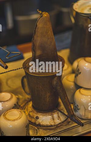 Eine glänzende traditionelle Kupferkaffeekanne steht auf einem Herd neben einem Stapel Keramikbecher. Der Topf hat ein klassisches Design mit einem abgerundeten Körper und einem Lo Stockfoto