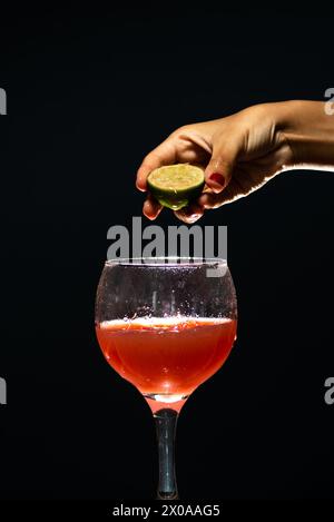 Eine Hand, die eine Zitronenscheibe über einem Glasbecher mit Guavasaft hält. Isoliert auf dunklem Hintergrund. Stockfoto