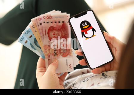 KIEW, UKRAINE - 1. APRIL 2024 Tencent QQ-Symbol auf Smartphone-Bildschirm und Geld in weiblicher Hand. IPhone-Display mit App-Logo und chinesischen Yuan-Geldscheine in Frauenhänden aus nächster Nähe Stockfoto