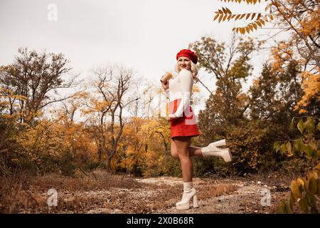 Eine Frau in rotem Rock und weißem Hemd posiert im Wald. Sie hält eine Tasse und hat einen roten Hut auf. Stockfoto