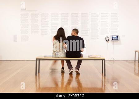 Ein Paar, das auf einer Bank in einer Museumsausstellung sitzt, lacht und auf die Wand mit Kunstwerken blickt Stockfoto