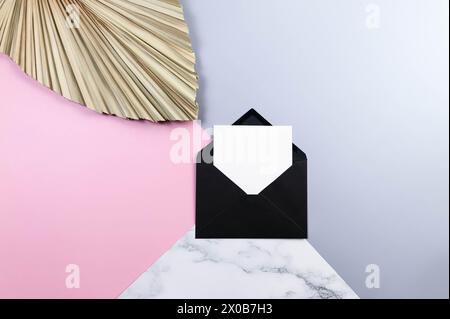Draufsicht auf schwarzen Umschlag, weiße Karte und getrocknete Palmblätter auf farbenfrohem Hintergrund. Blauer, rosafarbener und Marmorhintergrund. Pastellfarbene Tapete. Stockfoto