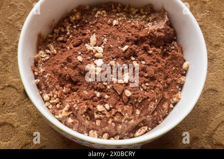 Blick von oben auf eine Schüssel mit cremigem, hausgemachtem Schokoladendessert, bestreut mit Mandeln und Nüssen und mit Kakaopulver auf einem Samtstuhl Stockfoto