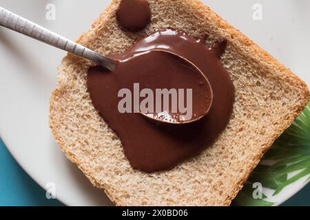 Blick von oben auf den wunderbaren Moment eines Löffels, der mit Schokoladenaufstrich überflutet wird, der auf ein Stück frisches Vollkorn-bre getränkt wird Stockfoto