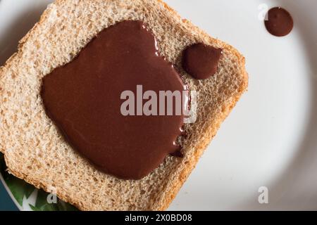 Blick von oben auf ein Stück Vollbrot und seine raue Textur mit einer glänzenden Haselnusscreme aus dunkler Schokolade, die auf einer weißen Platte platziert ist. Süße Stockfoto