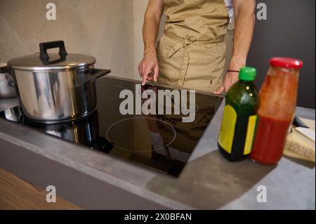 Nahaufnahme Mann in der Schürze des Küchenchefs, Hand auf schwarzer Glasoberfläche eines Elektro- oder Induktionsofens, Einschalten, Programm während des Kochens einstellen Stockfoto